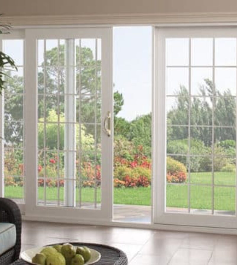 WHite 3-panel sliding patio door with white colonial grids and matching hardware. This is the inside of a livingroom with hardwood floors and a neutral color wall paint. The background is a nice, sunny day.